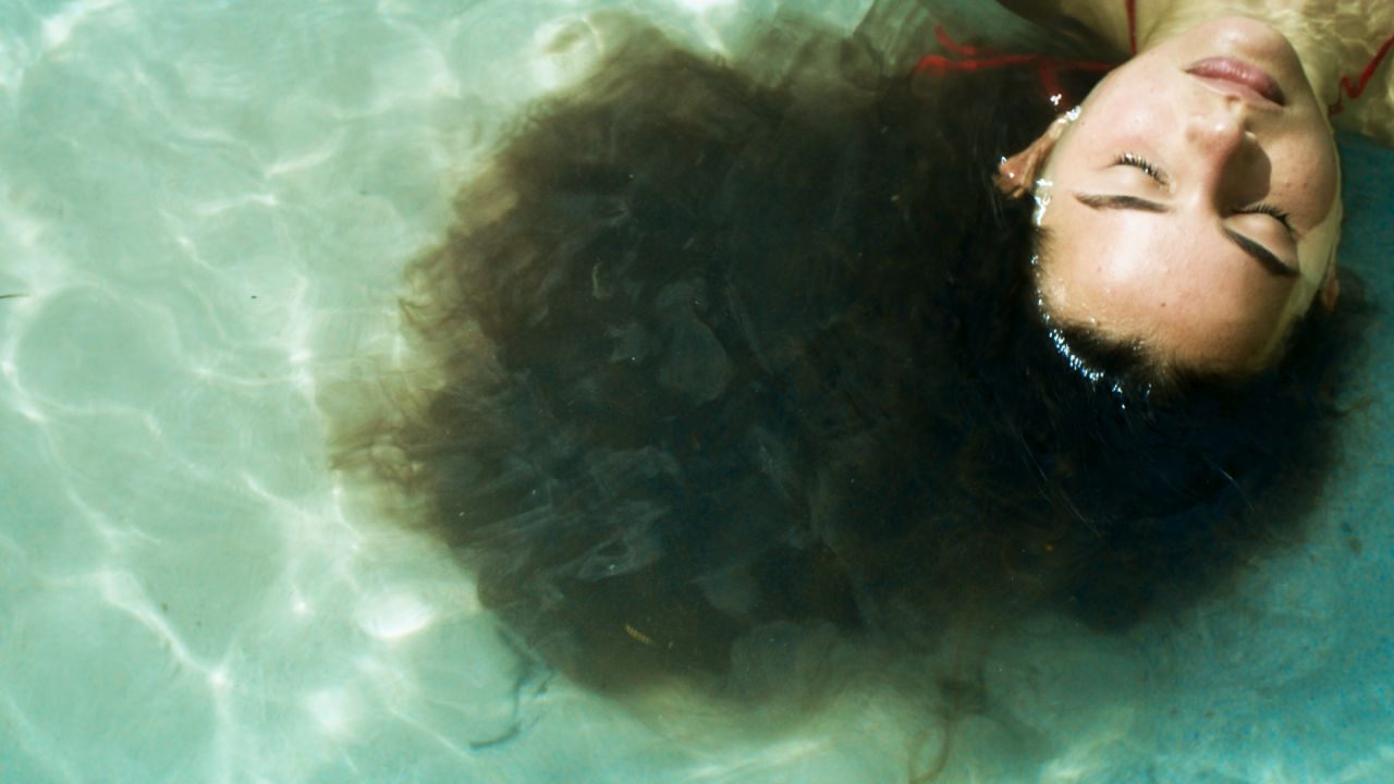 Person floating in clear water with eyes closed, hair fanned out in the water, and sunlight reflecting on the surface.