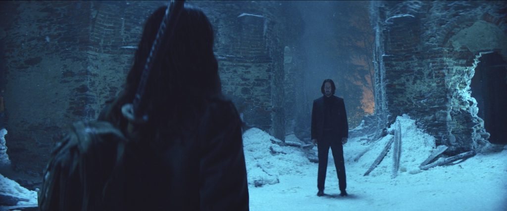 Two people face each other in a snow-covered, ruined stone building, with one seen from behind and the other standing ahead in the center.