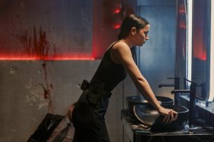 A woman in black stands at a sink in a dimly lit bathroom, appearing tense, with a person slumped on the floor in the background and blood stains on the wall.