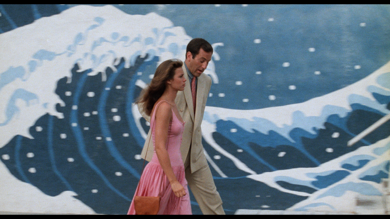 A woman in a pink dress and a man in a light suit walk past a mural of ocean waves.