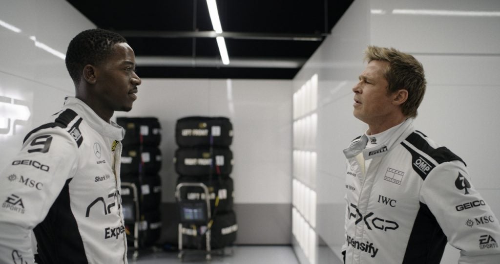 Two race car drivers in white racing suits stand facing each other in a garage with stacks of tires and equipment in the background.