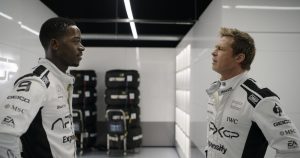 Two race car drivers in white racing suits stand facing each other in a garage with stacks of tires and equipment in the background.