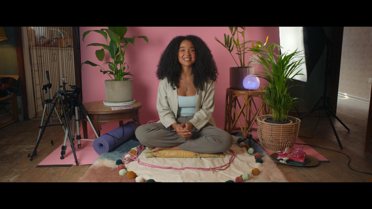 A woman sits cross-legged on a mat, surrounded by plants, a yoga mat, and camera equipment, against a pink backdrop in a home studio setting.