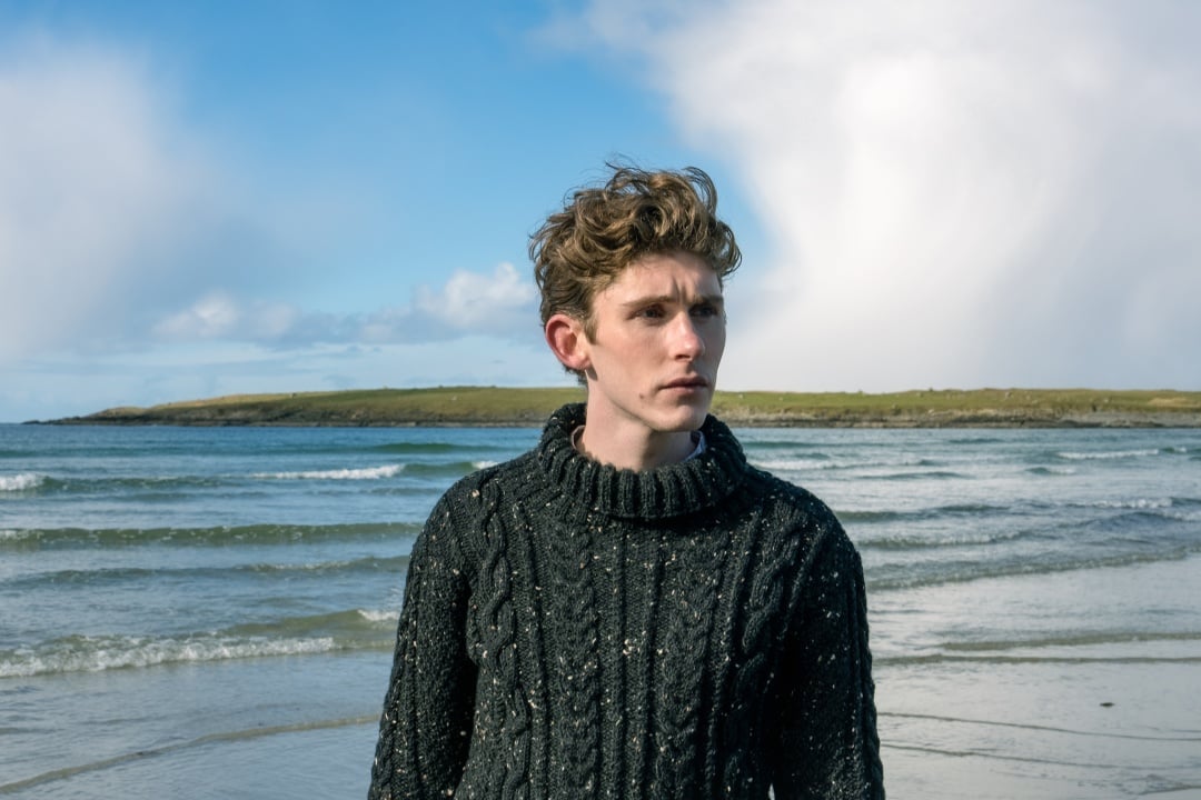 A young man with curly hair wearing a dark sweater stands on a beach with gentle waves and an overcast sky in the background.