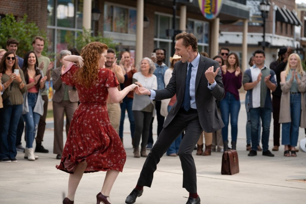 A man in a suit and a woman in a red dress dance together on a city sidewalk as a crowd of people watches and applauds.
