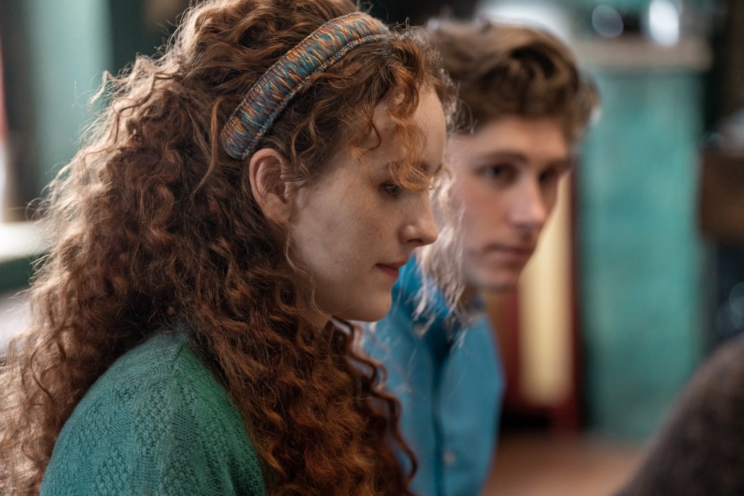 A woman with curly red hair and a headband is seated next to a man with wavy brown hair. Both are indoors and appear to be focused on something out of frame.