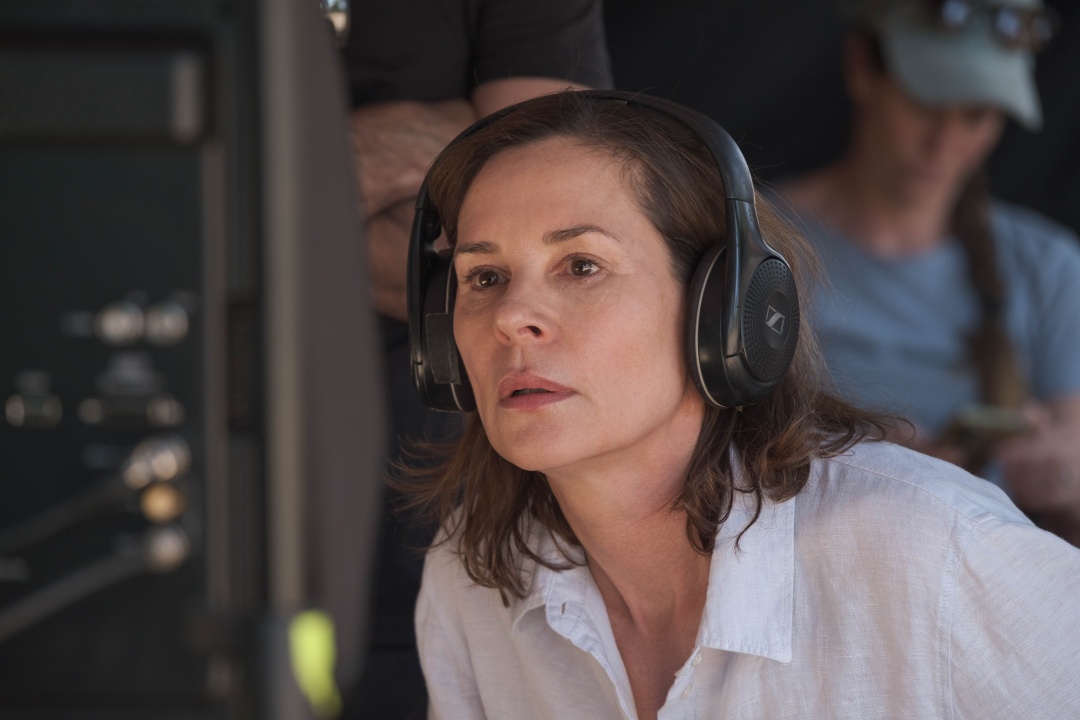 A woman wearing headphones and a white shirt closely observes a monitor, with another person blurred in the background.