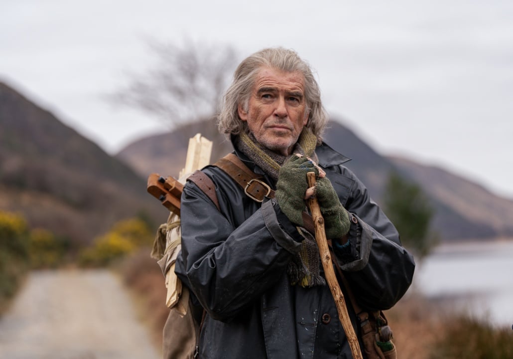 Older man with gray hair stands outdoors on a dirt path, holding a walking stick and wearing a coat, gloves, and scarf, with a backpack and mountains in the background.