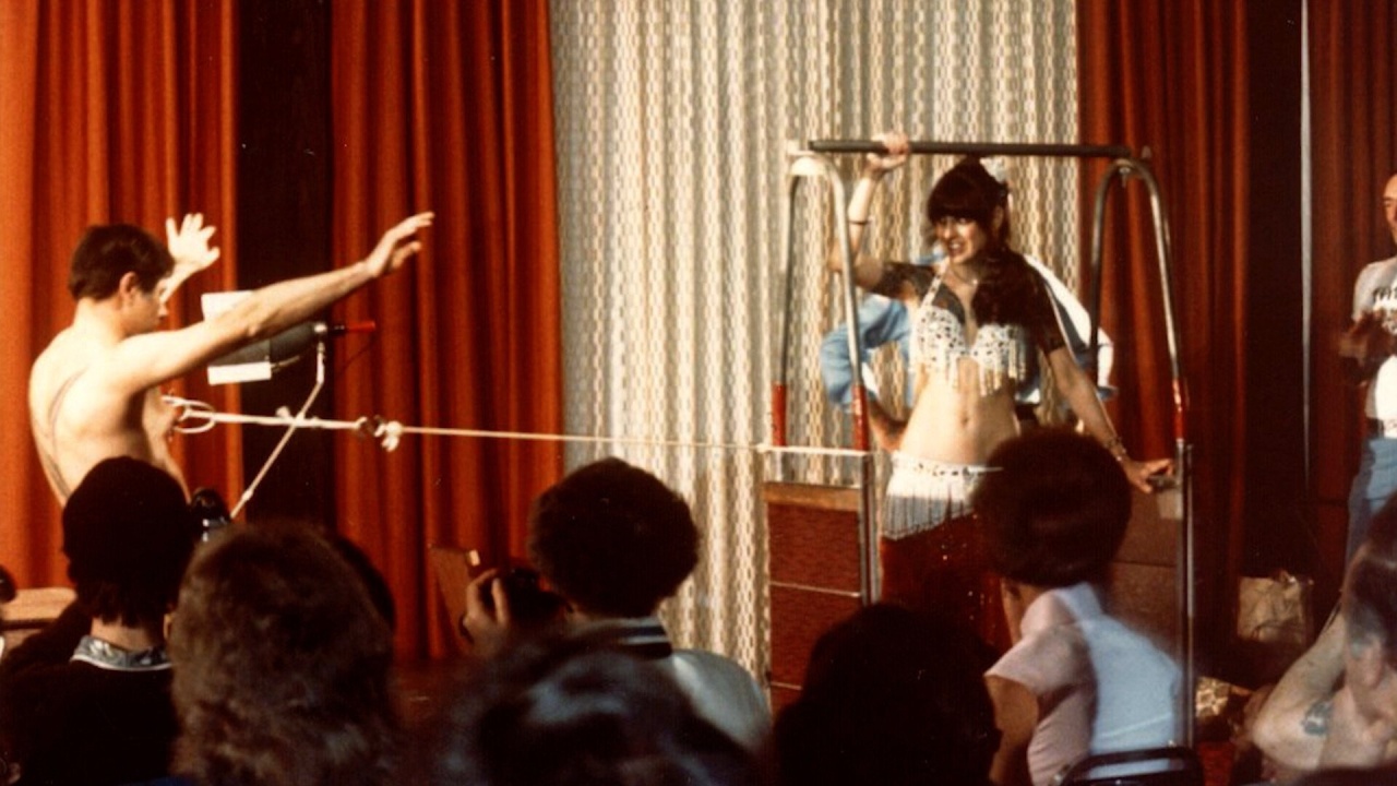 A man with a bow and arrow aims at a woman in a belly dancing costume standing behind a metal frame, while an audience watches indoors.
