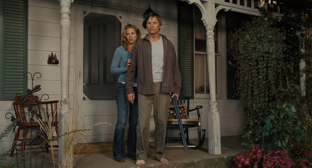 A man and a woman stand on the porch of a house, looking ahead with concerned expressions; the porch has a rocking chair and potted plants.
