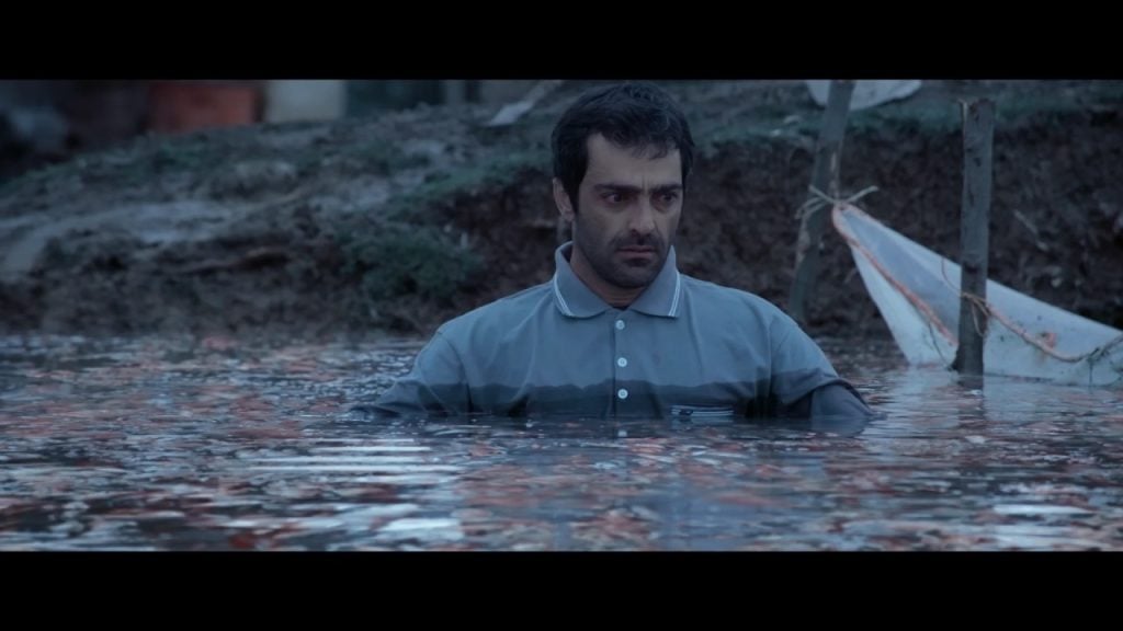 A man in a grey shirt stands chest-deep in murky water outdoors, looking serious, with muddy ground and a warning sign visible in the background.