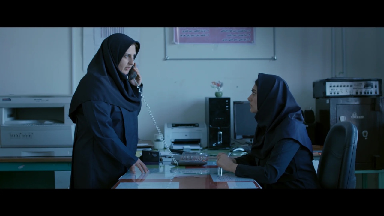Two women in dark clothing and headscarves are in an office; one is sitting at a desk using a computer, the other stands, speaking on a corded phone.