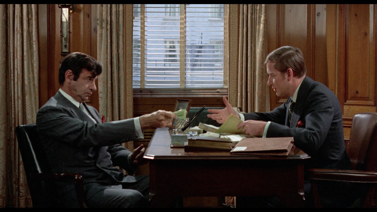 Two men in suits sit across from each other at a wooden desk in an office, exchanging documents, with files and papers spread out on the desk.