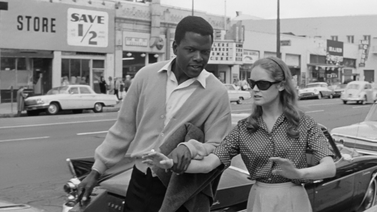 A man and a woman talk on a city sidewalk next to parked cars; storefronts and signs are visible in the background.