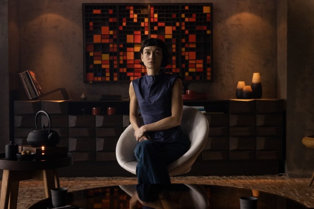 A person sits in a modern chair in a dimly lit room with geometric wall art, pottery, and a tea set on a low table.