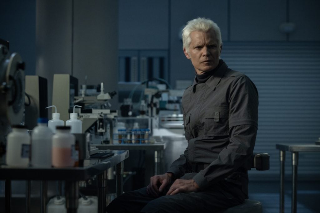A man with white hair in gray clothing sits in a modern laboratory, surrounded by scientific equipment and containers on metal tables.