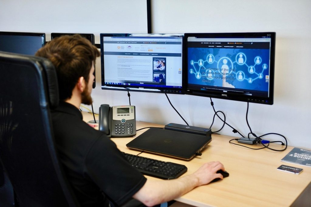 A person sits at a desk using a computer with dual monitors; a phone, closed laptop, and book are also on the desk.