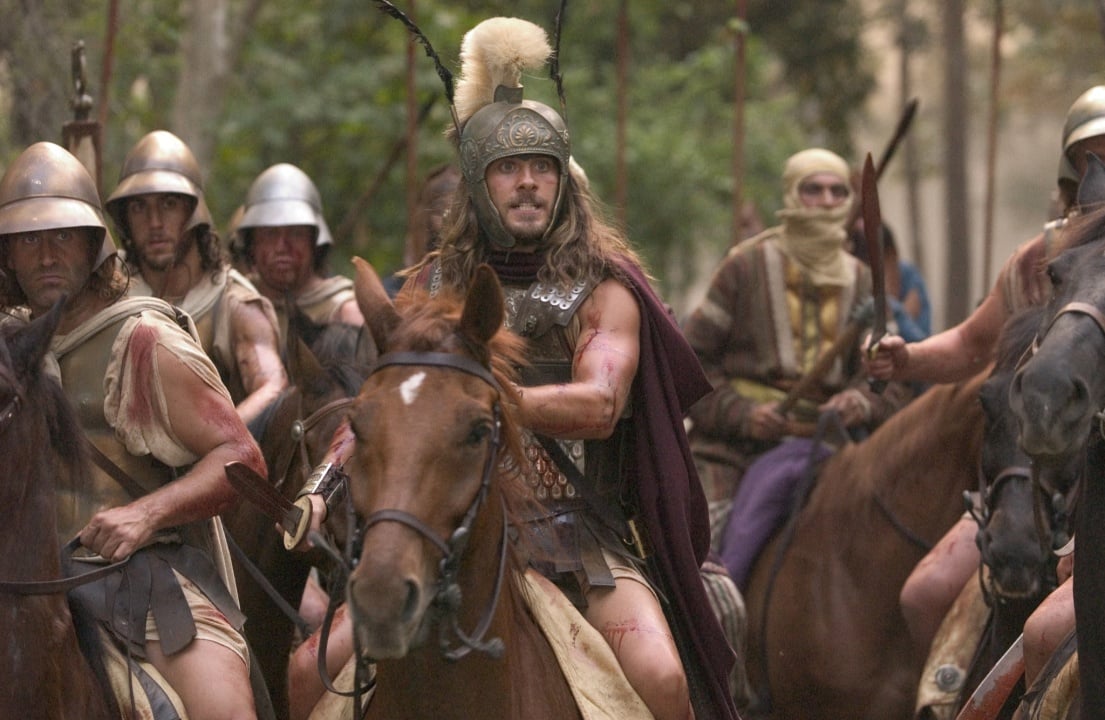Armored soldiers on horseback, led by a man in a helmet with a plume, move through a forested area, carrying spears and shields.