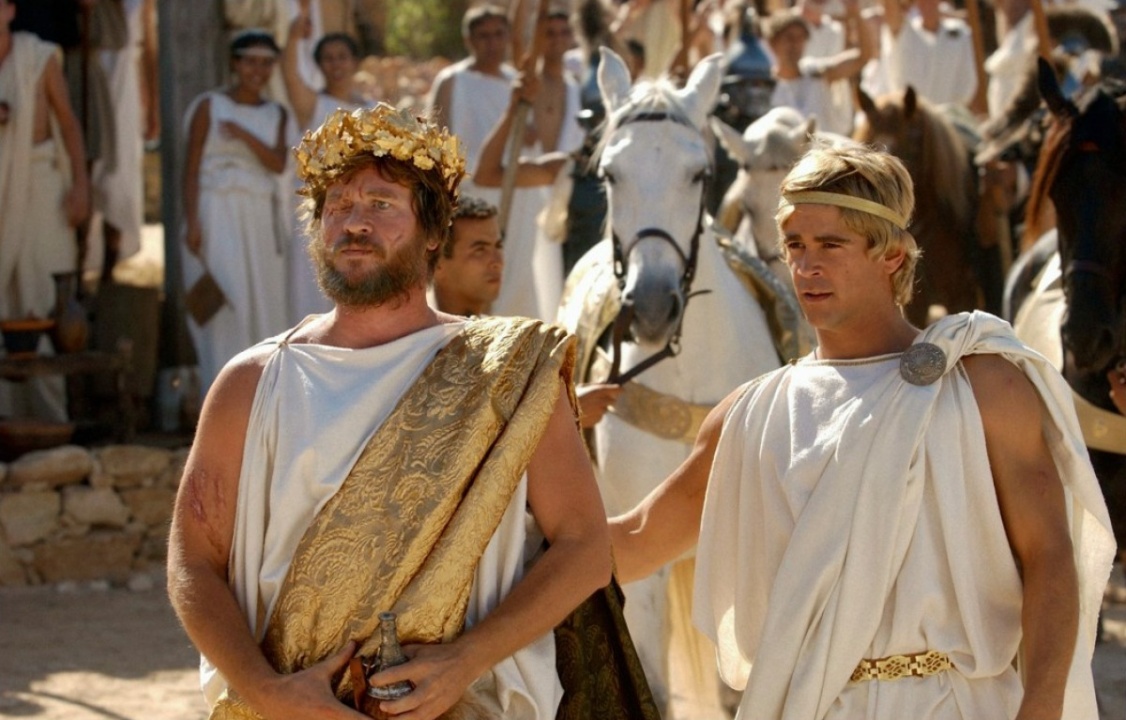 Two men in ancient Greek-style clothing and gold headpieces stand in front of white horses, surrounded by a crowd in similar attire at an outdoor gathering.