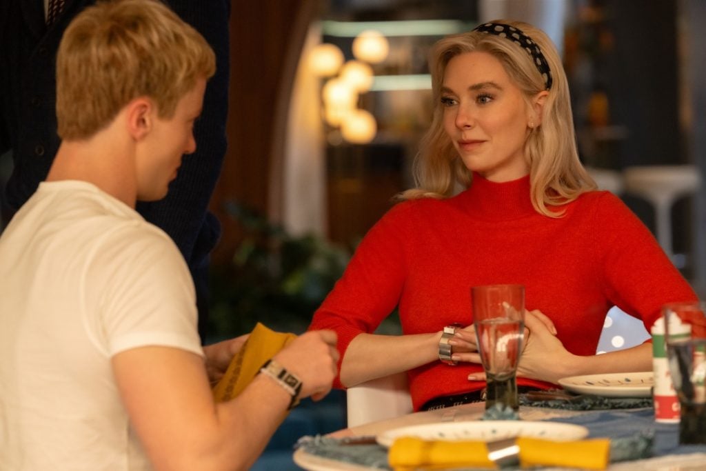 A woman in a red sweater sits at a table with a man in a white t-shirt; they appear to be having a conversation over a meal.
