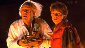 Two people stand side by side at night, one holding a device with wires and dials. Both look ahead with surprised expressions, illuminated by an orange light.