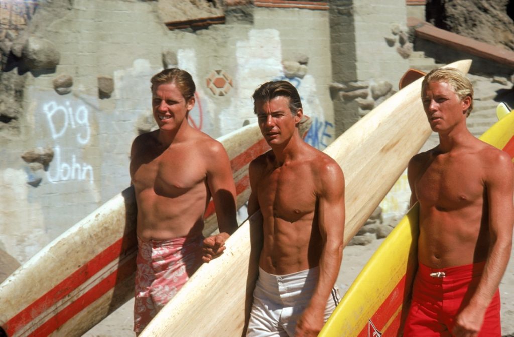 Three young men wearing swim trunks carry long surfboards while walking on a sandy beach, with graffiti and rocks visible on a weathered wall behind them.