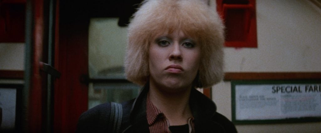 A person with blond, fluffy hair and a serious expression stands in front of a wall with signage and a window in the background.