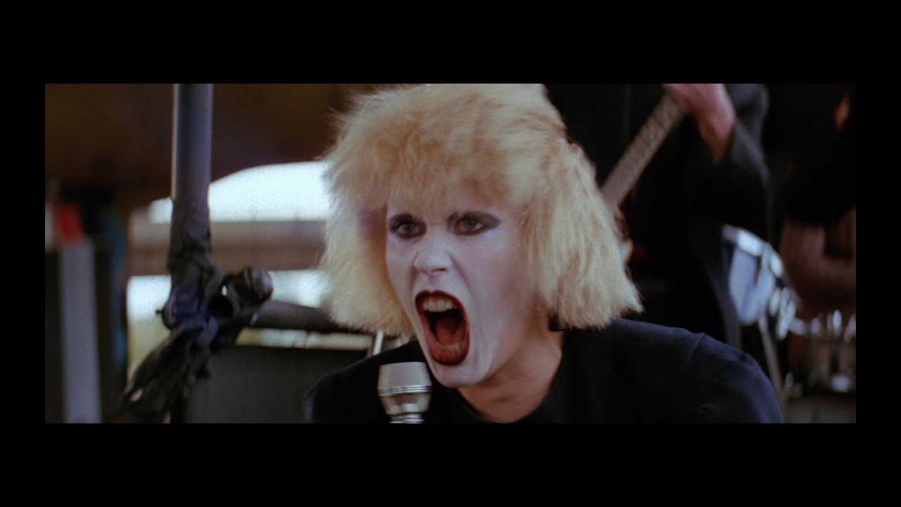 A person with pale skin, bleached blond hair, and heavy dark makeup shouts into a microphone while performing on stage.