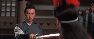A man in traditional attire points a sword at someone wearing red, while several people watch in the background inside a wooden room.