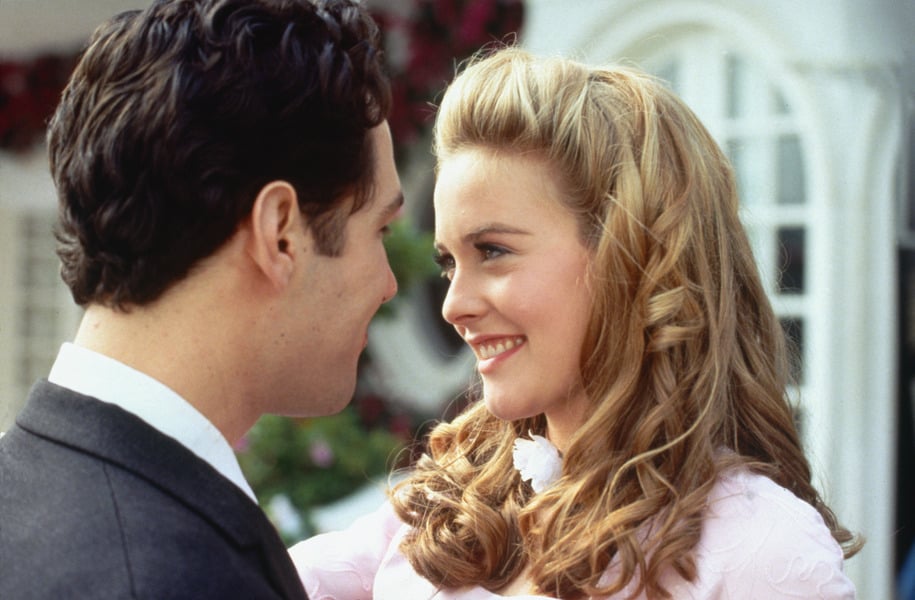 A young woman with long blonde hair smiles at a young man in a suit as they stand close together outside a building with arched windows.