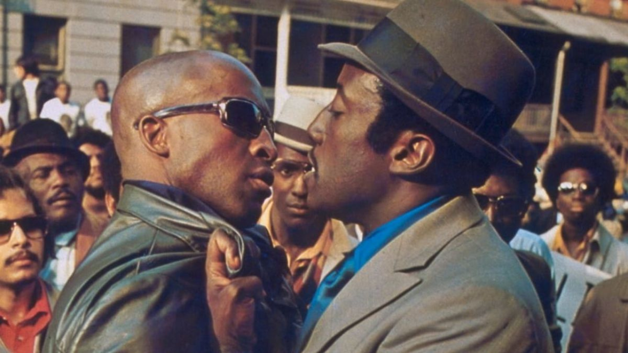 Two men confront each other closely in an intense face-off, with one gripping the other's jacket, while a group of people watch in the background.