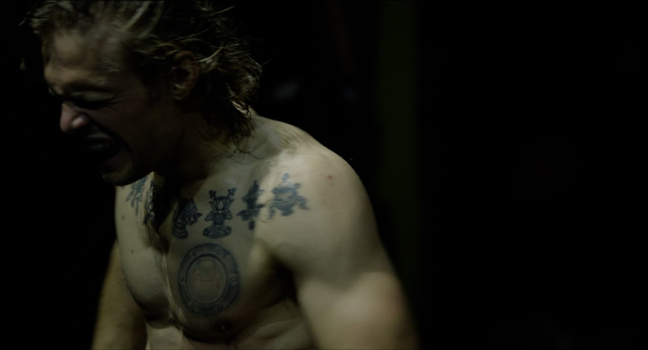 A shirtless person with tattoos on their chest is standing in a dimly lit area, appearing to be exerting effort or in distress.