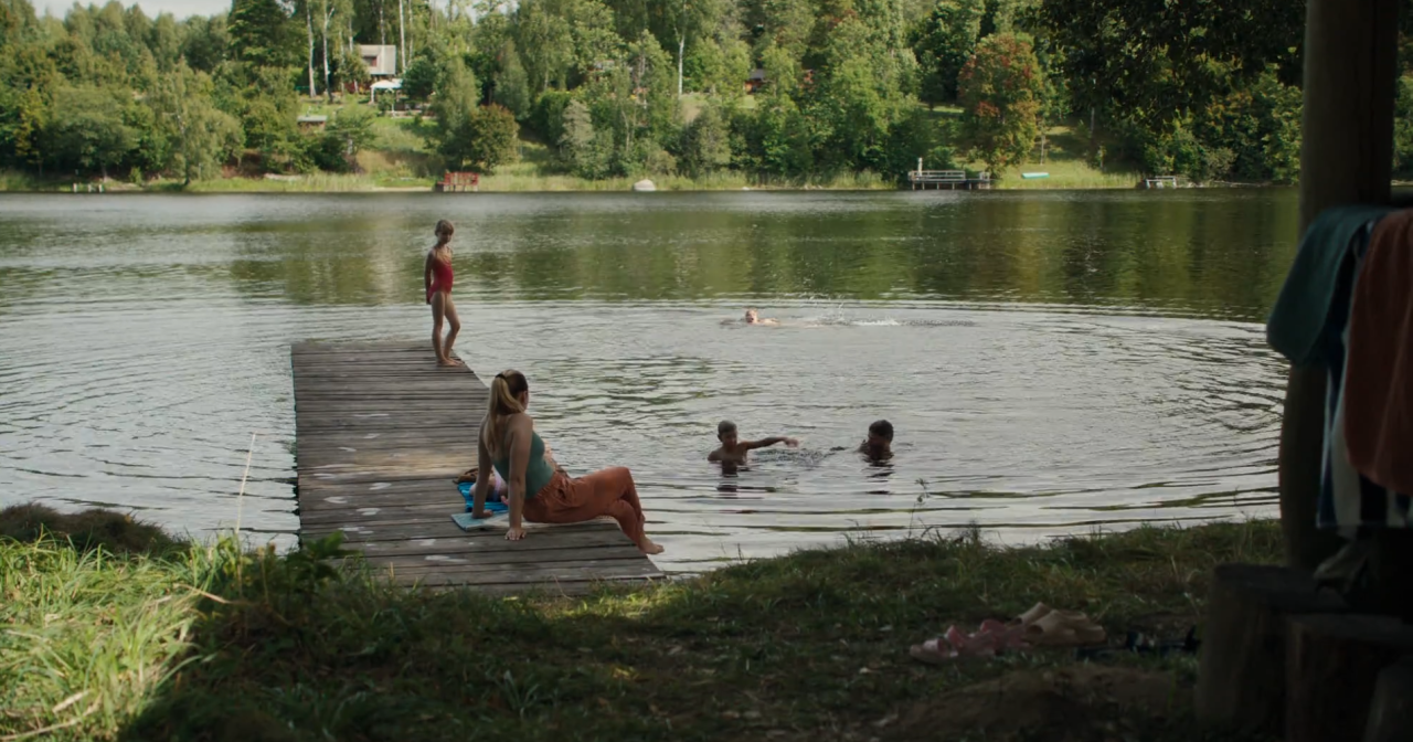 People are swimming and relaxing by a lakeside dock surrounded by trees, with one person sitting on the dock and others in the water.