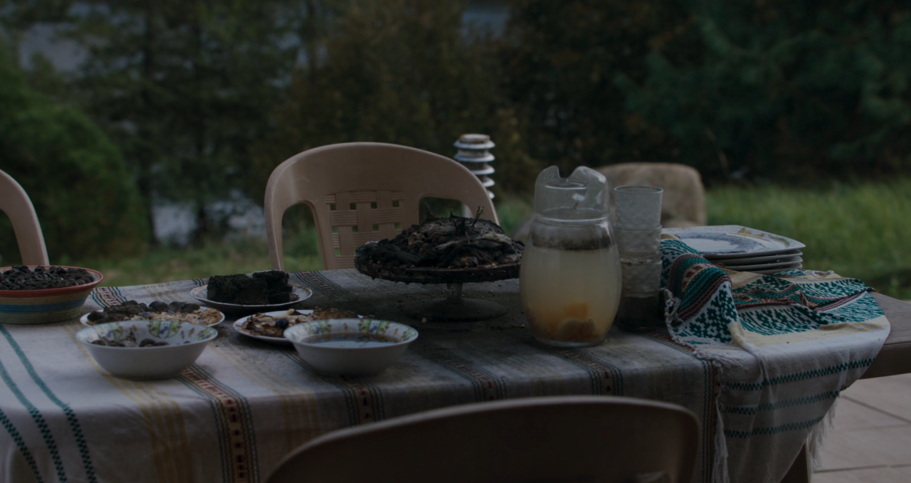 A table outdoors with empty chairs, plates of food, a bowl, a pitcher of drink, and a folded patterned cloth on a checkered tablecloth. Trees are visible in the background.