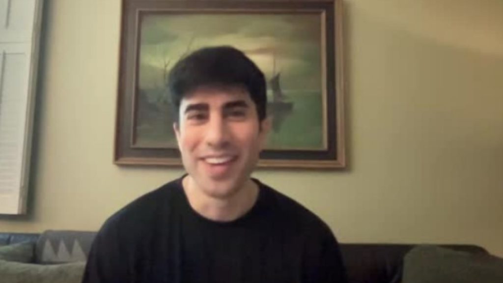 A person with short dark hair, wearing a black shirt, sits on a couch and smiles. A framed painting of a ship hangs on the wall in the background.