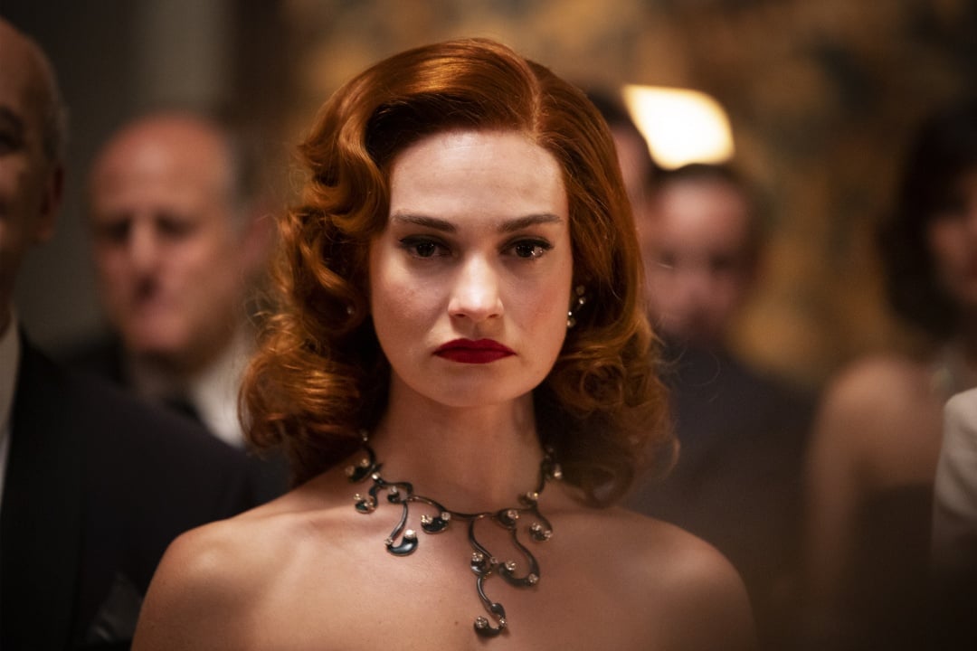 A woman with wavy red hair and bold red lipstick wears a black statement necklace, standing in a formal setting with blurred people in the background.