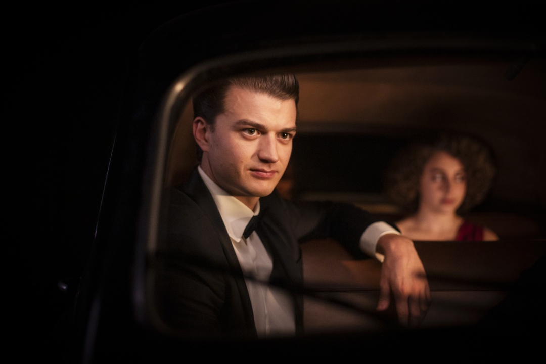 A man in a tuxedo sits in the front seat of a car at night, looking forward, with a woman in a red dress seated in the back.