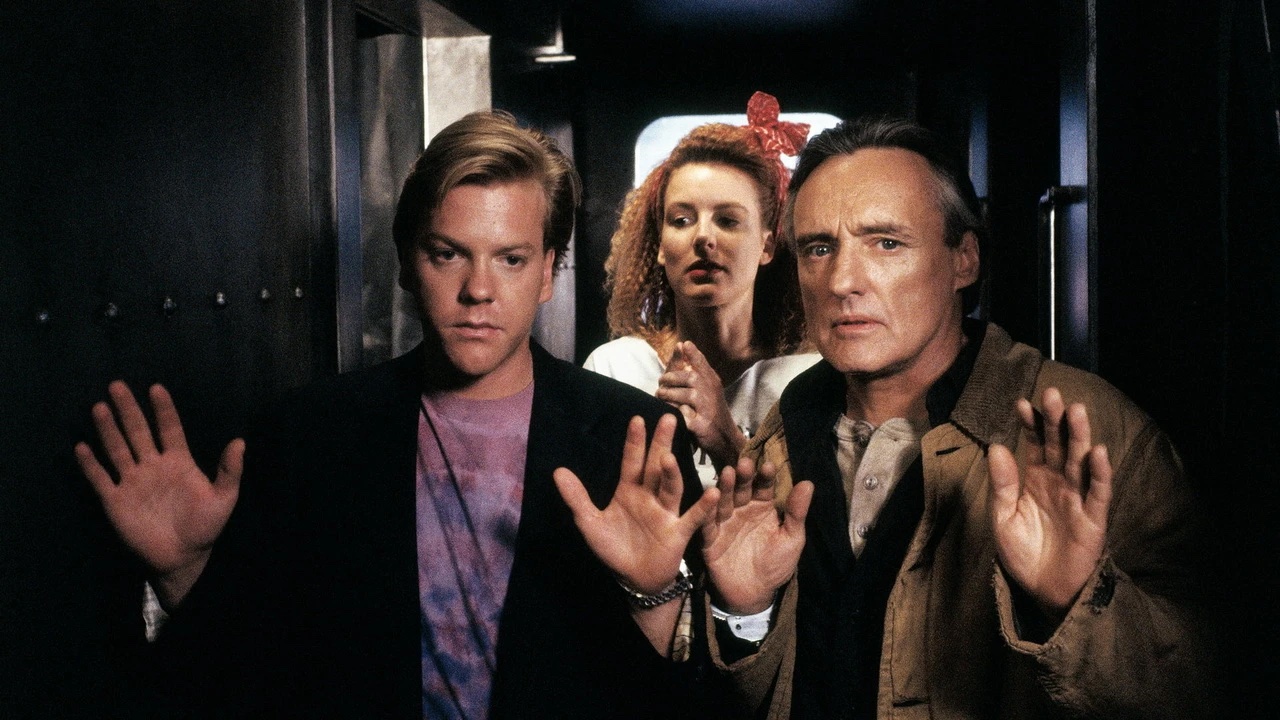 Two men with raised hands and a woman behind them stand in a dimly lit hallway, appearing concerned or cautious.