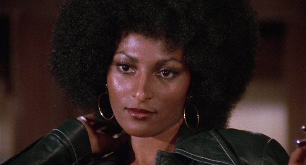 A woman with a large afro hairstyle looks forward, wearing hoop earrings and a dark jacket, with a neutral expression.