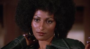 A woman with a large afro hairstyle looks forward, wearing hoop earrings and a dark jacket, with a neutral expression.