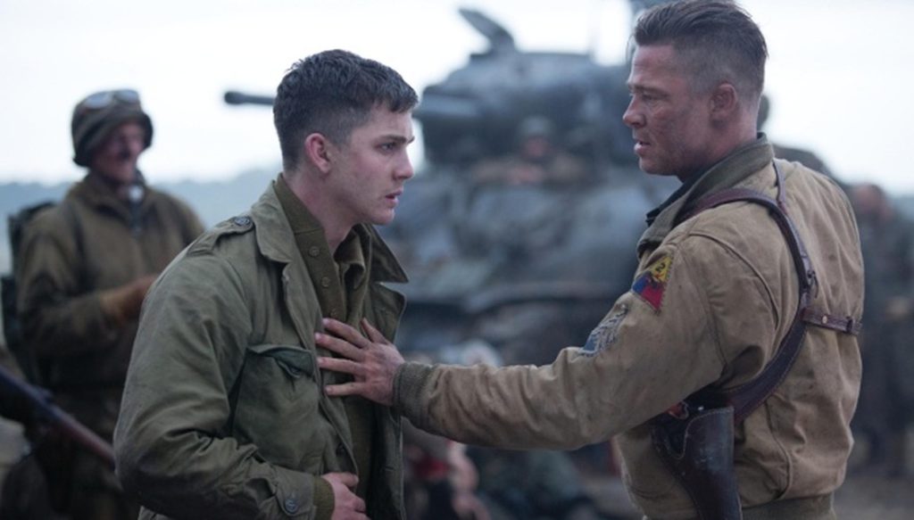 Two soldiers in military uniforms stand facing each other, one placing a hand on the other's chest, with a tank and other soldiers in the blurred background.