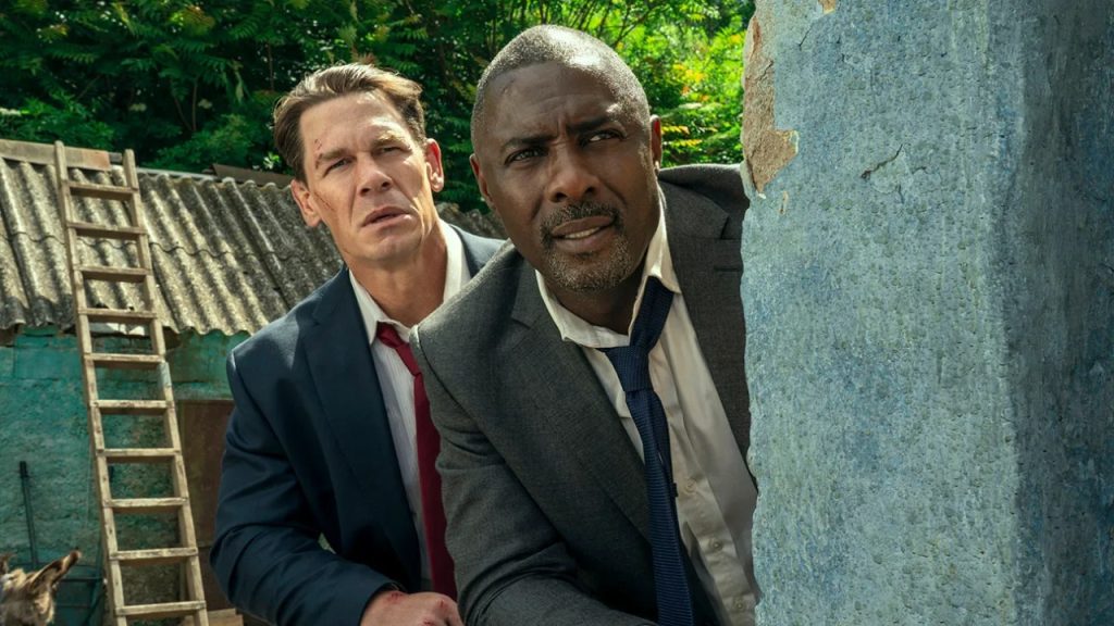 Two men in suits, one with a red tie and one with a blue tie, crouch behind a wall, looking intently ahead in an outdoor setting with greenery behind them.