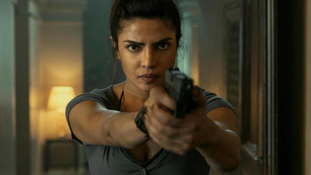 A woman with tied-back hair points a handgun forward with both hands, standing indoors in a dimly lit hallway.