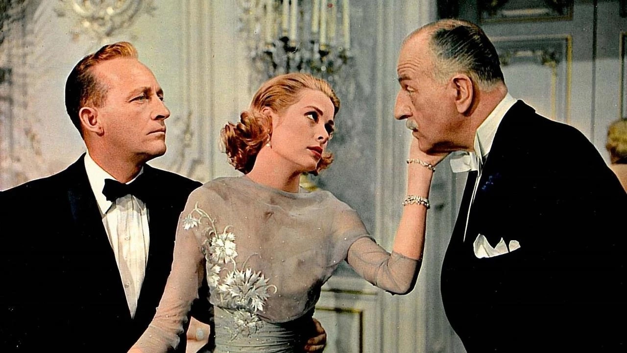 A woman in an elegant dress stands between two men in tuxedos; she gently holds the chin of the older man while the younger man watches.