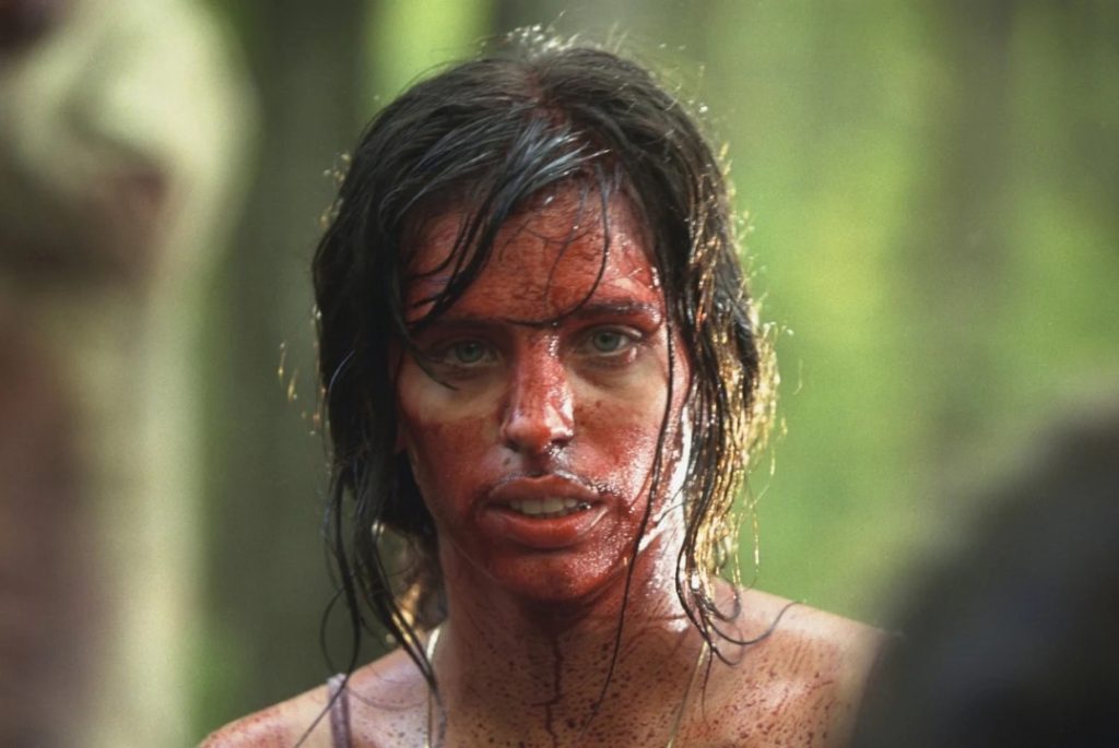 A person with long dark hair and bare shoulders stands in a forest, their face and upper body covered in blood, looking directly at the camera.