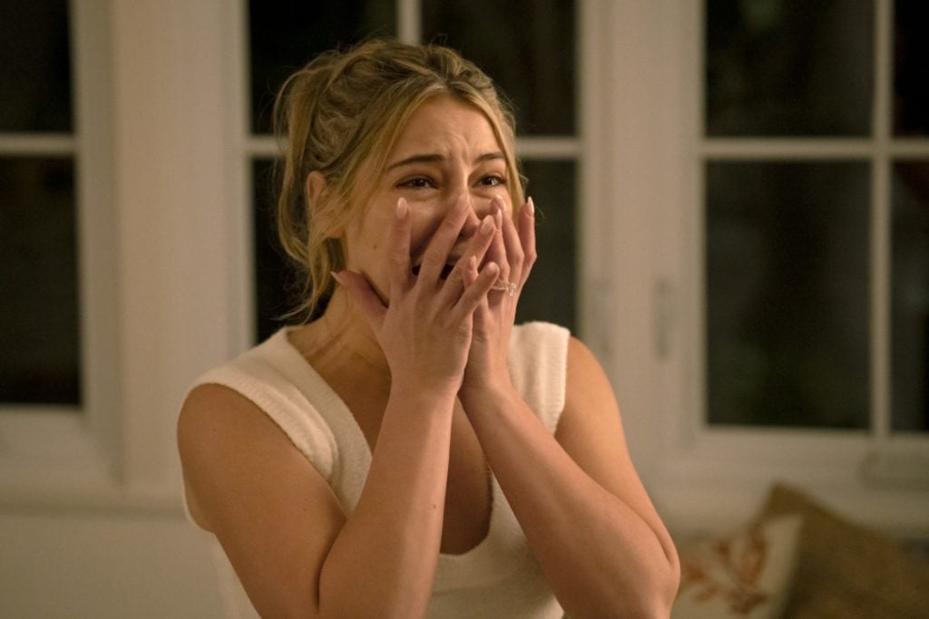 A woman with light hair in a ponytail covers her mouth with both hands, appearing shocked or distressed, standing indoors in front of a window.