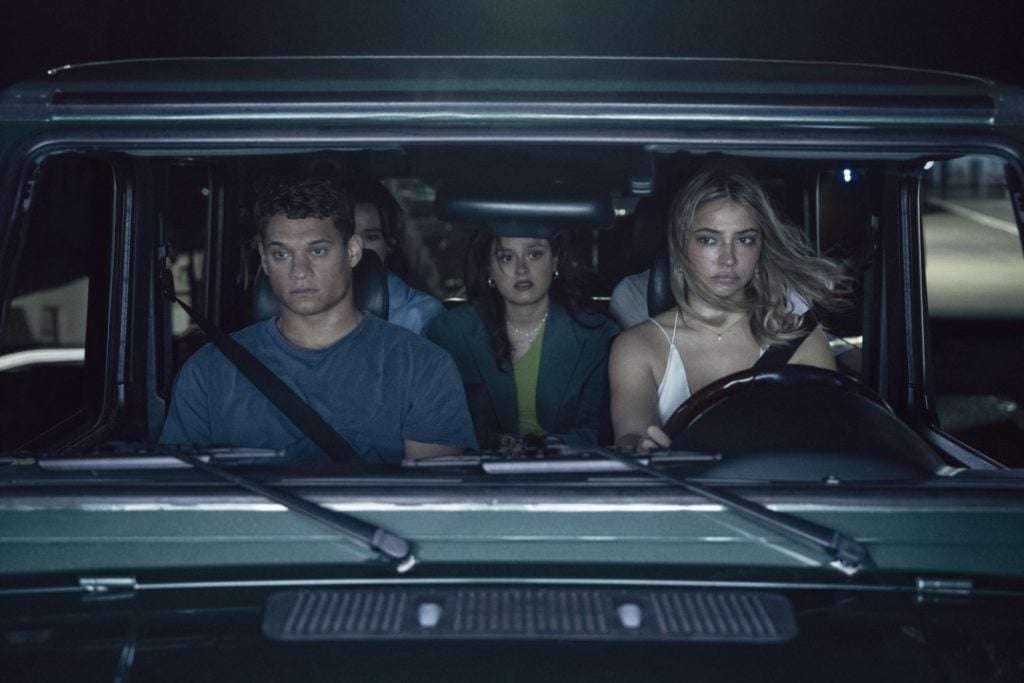 Four young adults sit in a car at night, with a woman driving and the others looking ahead with serious expressions.