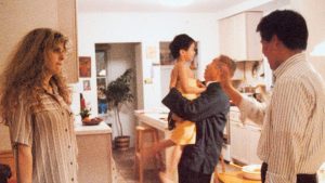 Four people are in a kitchen; a woman stands on the left, while a man lifts a child and another man stands nearby, all appearing to interact together.