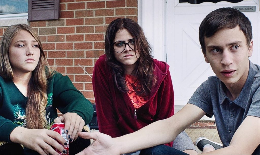 Three teenagers sit outside in front of a brick wall and white door; one holds a soda can, another wears glasses, and the third gestures with his hand while talking.