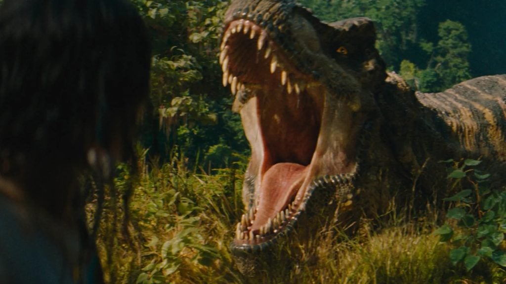 A large dinosaur with its mouth wide open faces a person in a dense, green forest.